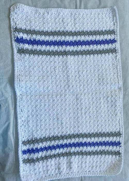 A crochet white towel dish towel with striped grey and blue at the ends displayed on a white surface
